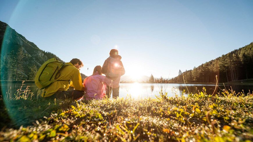 Eine kleine Familie sitzt bei Sonnenschein an einem See.