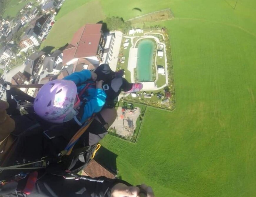 Blick von oben auf eine Person mit Kind beim Paragliding über eine grüne Landschaft und ein Resort mit Pool.