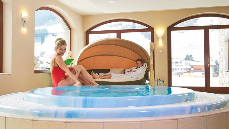 Eine Frau im roten Badeanzug genießt den Whirlpool, während ein Mann in einem bequemen Liegestuhl mit Baldachin im Hintergrund entspannt.