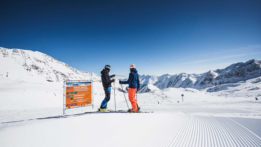 Zwei Skifahrer stehen auf der frischen Piste und unterhalten sich.