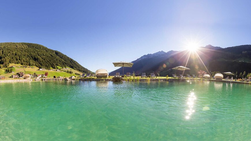 Der türkisfarbene Naturbadeteich mit Blick auf die Berglandschaft und strahlenden Sonnenschein