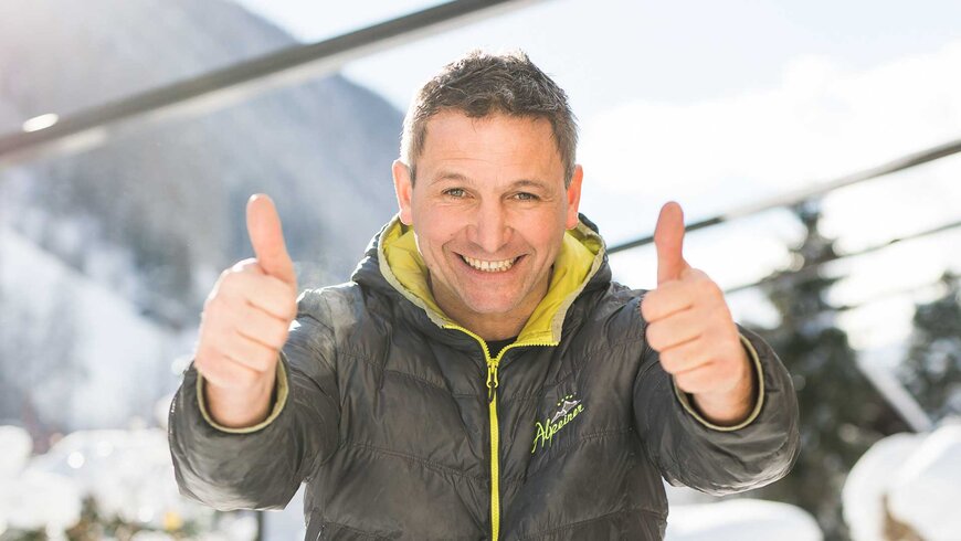 Ein Mann in schwarz-gelber Winterjacke lächelt und streckt beide Daumen nach oben in die Kamera.