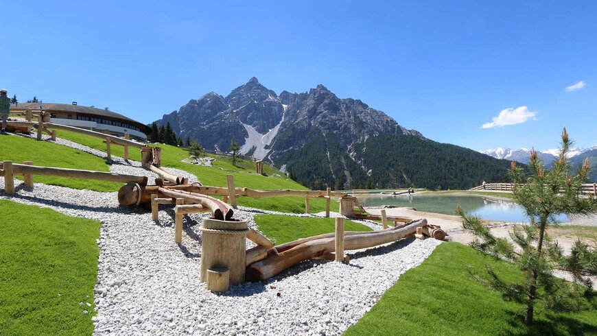 Ein Naturspielplatz mit Holzkonstruktionen und einem Teich inmitten der malerischen Berglandschaft Tirols.