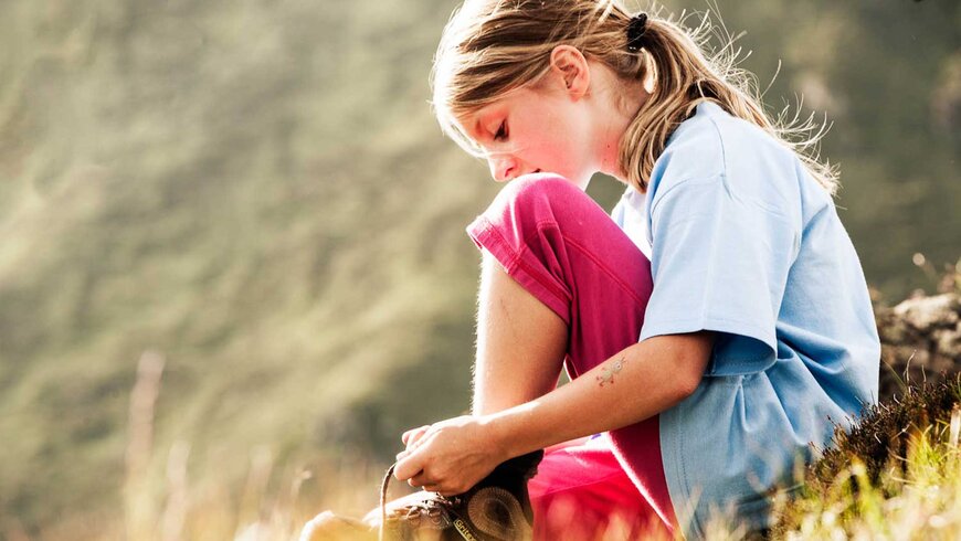 Ein Mädchen sitzt in der Natur und bindet ihre Schuhe während eines Sommerurlaubs mit der Familie in Tirol.