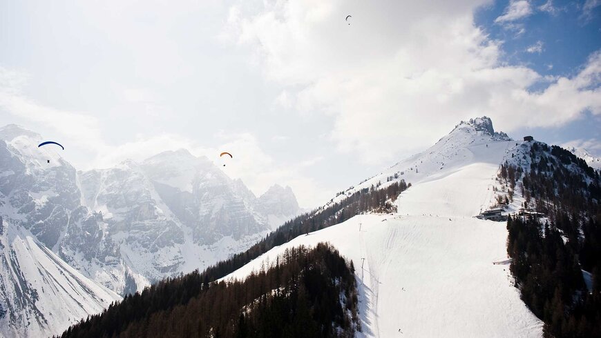 Paragleiter fliegen während des Aktivurlaub in Tirol über verschneiten Berggipfel und Skipisten bei sonnigem Wetter.