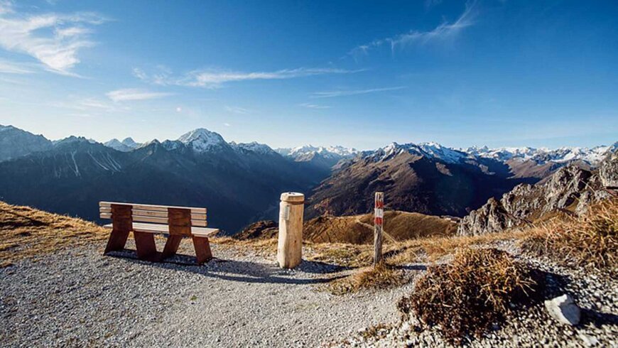 Holzbank auf einem Berggipfel mit atemberaubendem Blick auf eine Alpenlandschaft bei klarem Himmel.