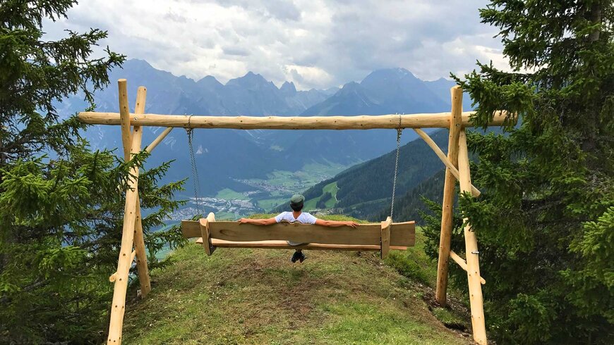 Ein Mann sitzt auf einer Holzschaukel und genießt den atemberaubenden Blick auf die Tiroler Alpenlandschaft während eines Sommerurlaubs in Tirol.