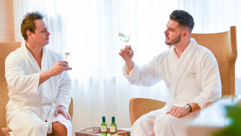Zwei Hotelgäste im Bademantel mit einem Glas Weißwein in der Hand im Wellnessbereich