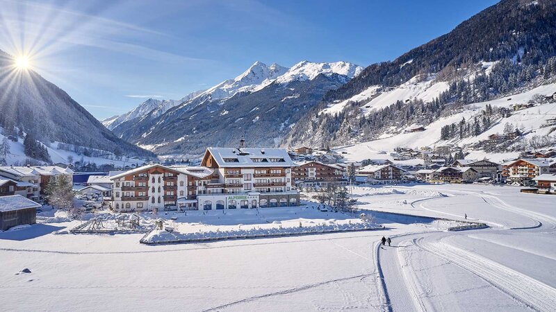Der Alpeiner Nature Resort inmitten eines malerischen Dorfs mit schneebedeckten Bergen und klarer Wintersonne im Hintergrund.