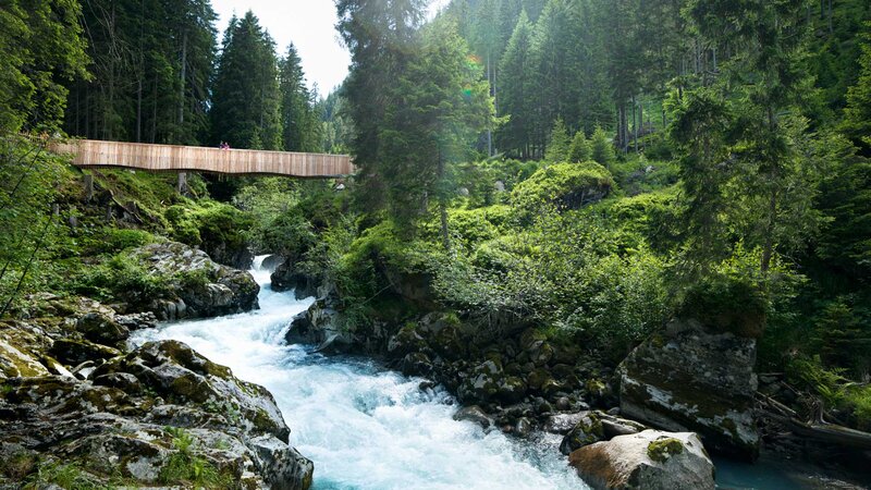 Ein rauschender Gebirgsbach fließt durch einen dichten Wald in Tirol, überquert von einer Holzbrücke.