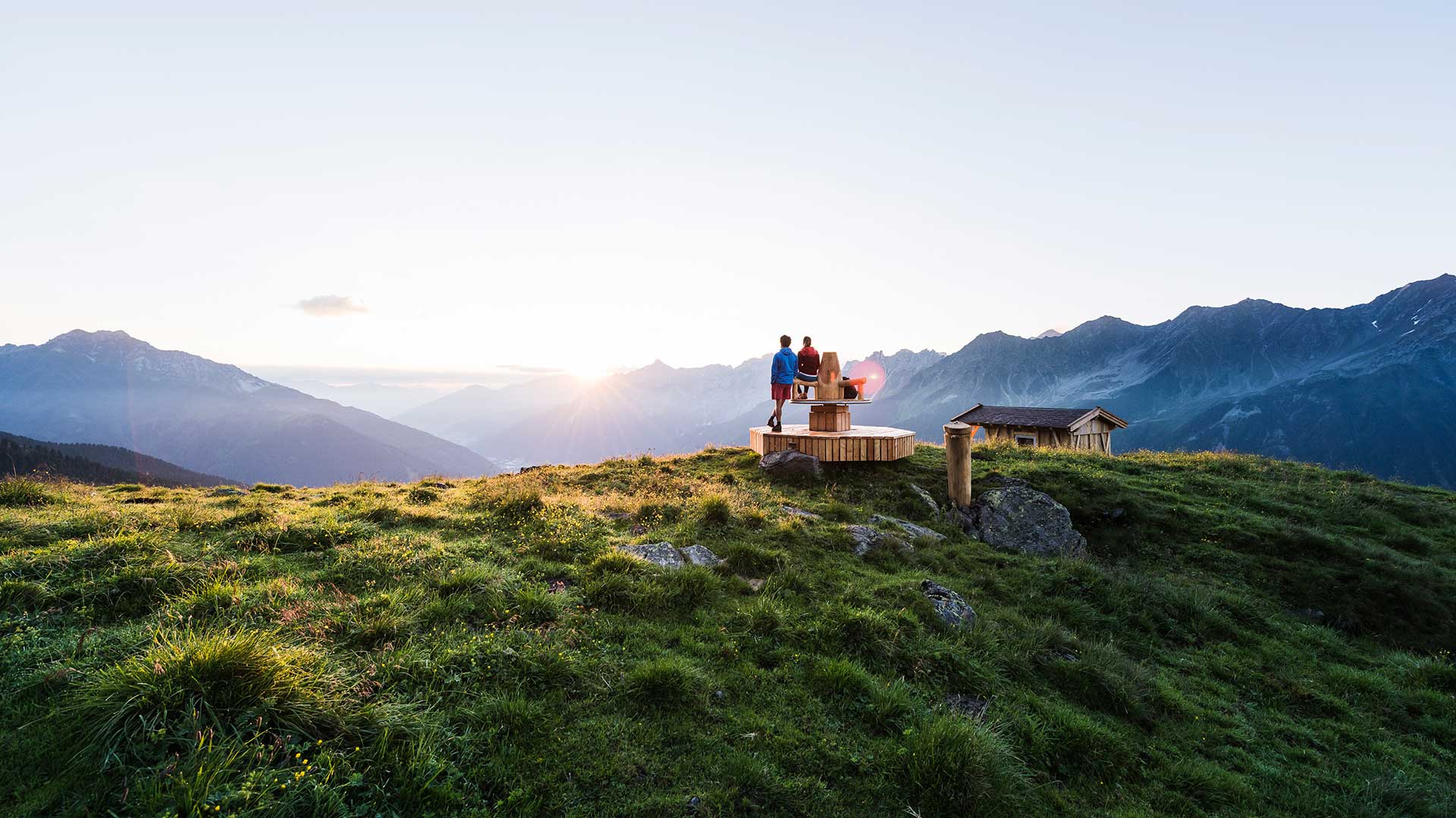 Zwei Menschen sitzen auf einer hölzernen Aussichtsplattform und genießen den Sonnenuntergang während ihres Sommerurlaubs in Tirol, mit einem weiten Blick auf die umliegenden Berge und Täler.