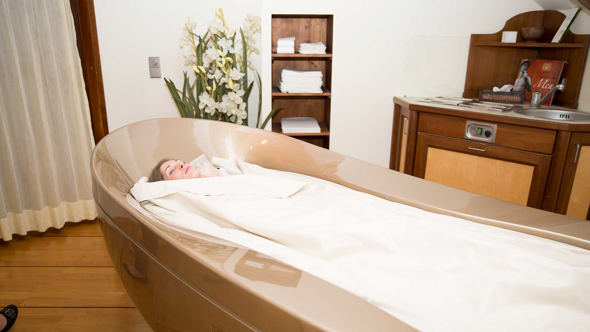 Eine Frau entspannt in einer Wellnessbehandlung, eingehüllt in ein wärmendes Tuch in einem modernen Spa-Raum.