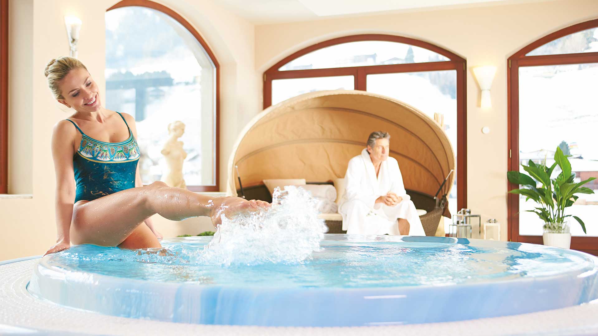 Eine Frau im Badeanzug sitzt Beim Wellness im Stubaital am Rand eines runden Whirlpools und spritzt spielerisch mit dem Wasser, während ein Mann in einem weißen Bademantel auf einer Liege im Hintergrund sitzt.