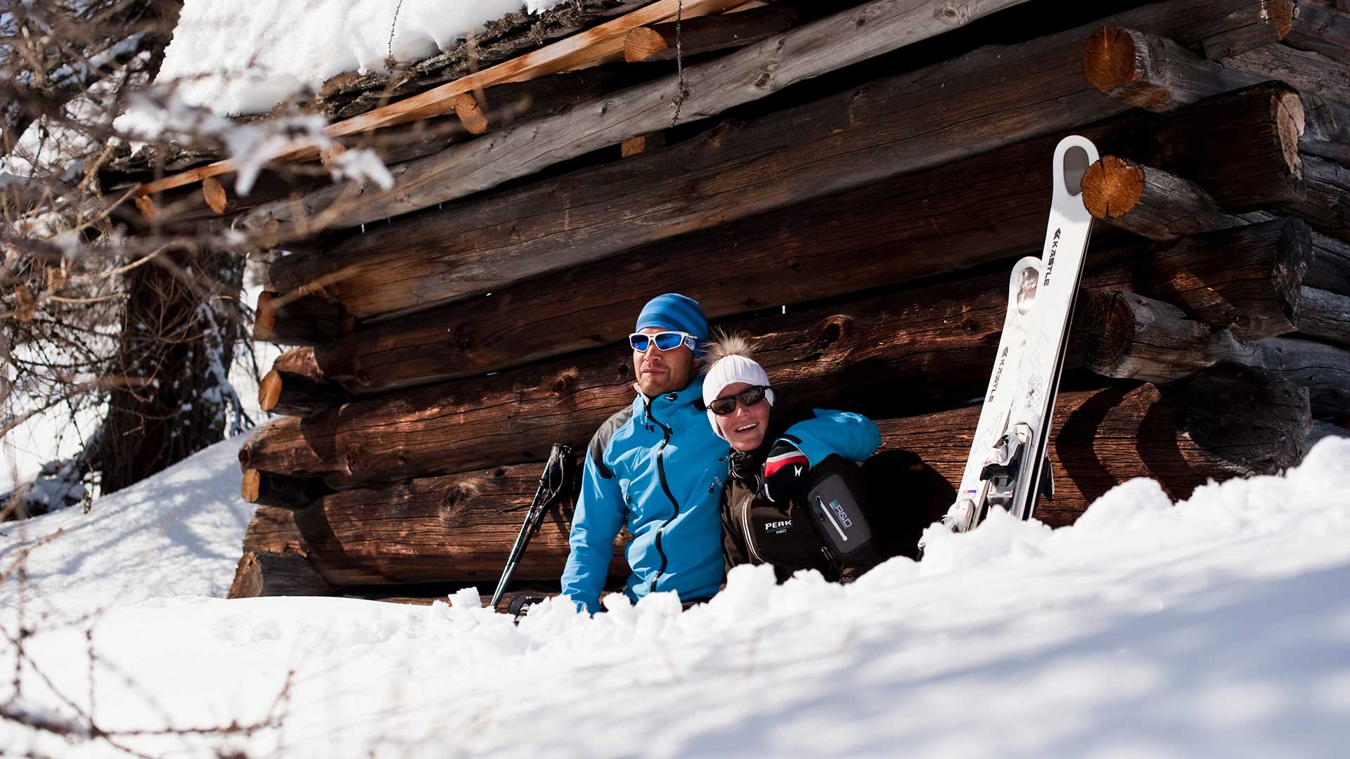 Ein Paar mit Skiern sitzt auf einer Holzbank in einer verschneiten Landschaft. 