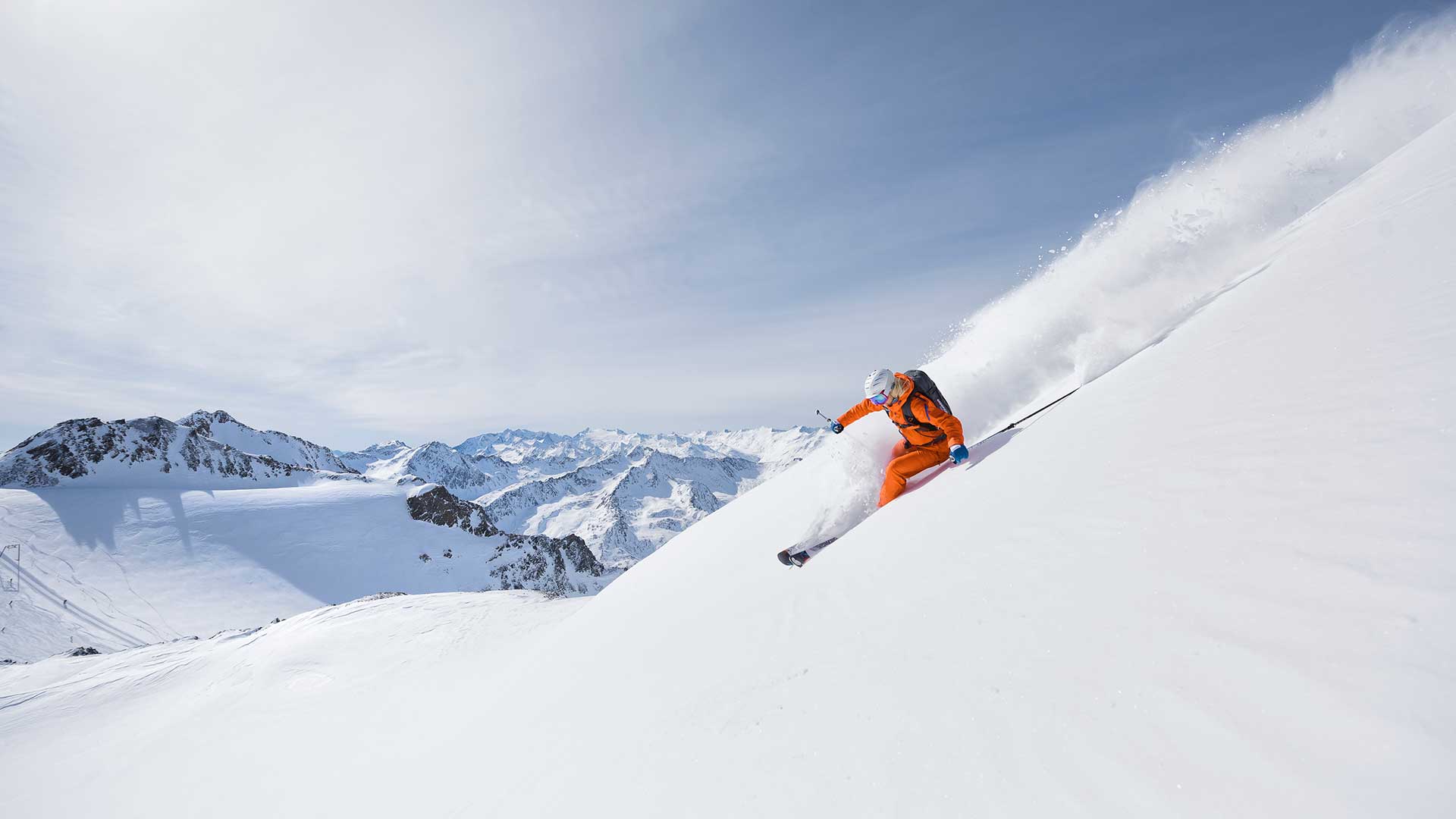 Skifahrer in orangem Anzug, der eine steile, schneebedeckte Piste im Stubaital hinabfährt, mit atemberaubendem Bergpanorama im Hintergrund.