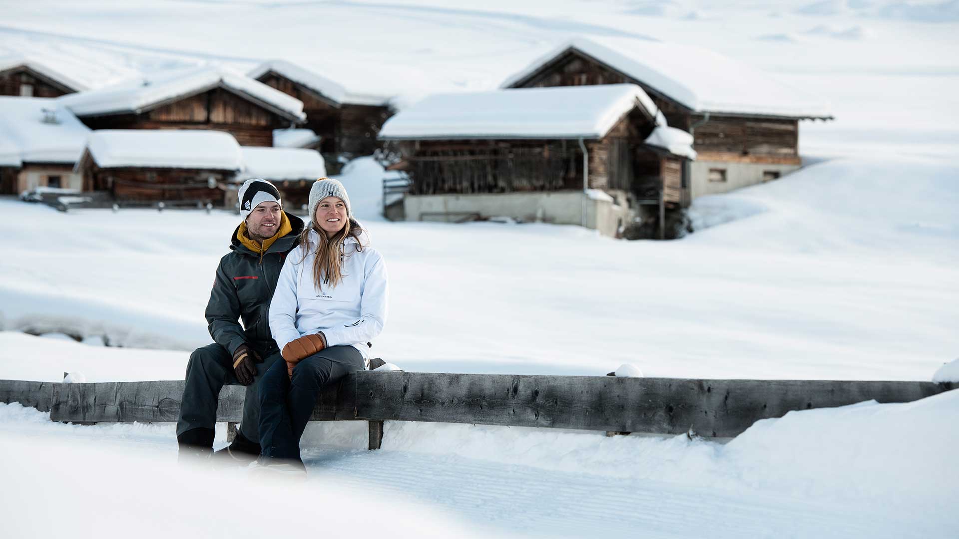 Paar in Winterkleidung sitzt während des Aktivurlaubs auf einer Bank vor traditionellen Holzhütten in einer verschneiten Landschaft in Tirol.